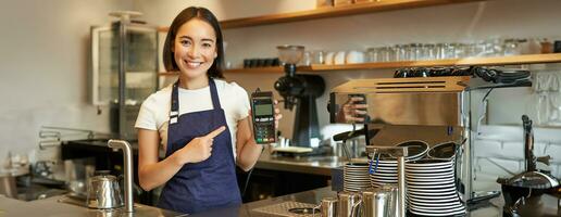 lächelnd asiatisch Mädchen Barista, Cafe Inhaber im Schürze, zeigen Karte Maschine, Zahlung Leser, nehmen kontaktlos Aufträge im ihr Kaffee Geschäft foto