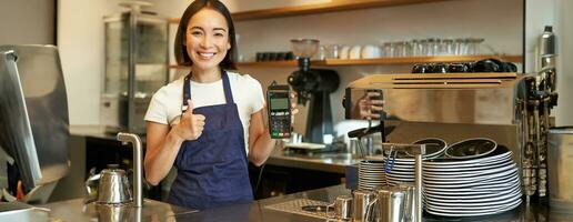 lächelnd asiatisch Mädchen Barista, Cafe Inhaber im Schürze, zeigen Karte Maschine, Zahlung Leser, nehmen kontaktlos Aufträge im ihr Kaffee Geschäft foto