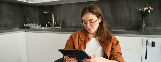 Porträt von Brünette Frau im Gläser, Sitzung im Küche beim heim, halten Digital Tablette, lesen auf Gadget foto