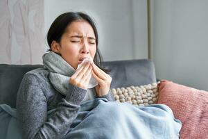 Bild von krank Koreanisch Frau beim heim, bedeckt im warm Kleider und Schal, Gefühl krank, fangen ein kalt und Sitzung auf Sofa Niesen foto