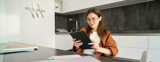 Porträt von jung Brünette Frau im Gläser, Sitzung im ihr Küche mit Digital Tablette, lesen auf Gerät, Aufpassen Videos online, Arbeiten auf Fernbedienung von ihr Zuhause foto