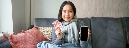 lächelnd Koreanisch Mädchen zeigt an Smartphone Bildschirm, Medikation im Hände, Gefühl krank und bleiben beim heim, mit online gp Arzt App während auf krank verlassen foto