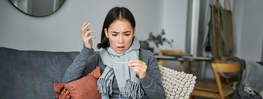 schockiert Frau sieht aus besorgt beim ihr Thermometer, fangen kalt, hat Fieber, Sitzung auf krank verlassen beim Zuhause foto