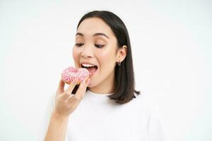 schließen oben von lächelnd zufrieden asiatisch Frau, isst glasiert Rosa Krapfen, genießt Essen lecker Donut, Weiß Studio Hintergrund foto