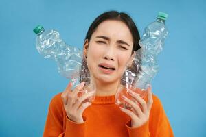 Porträt von Weinen asiatisch Frau mit zwei Plastik Flaschen, müde von Recycling, Sortierung Müll, Blau Hintergrund foto