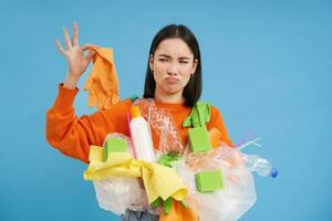 Koreanisch Frau sieht aus beim stinkend Latex Handschuh, hält Plastik Müll, reinigt Haus und recyceln, Blau Hintergrund foto