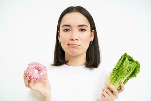 gesund sauber Entgiftung Essen Konzept. Vegetarier, vegan, roh Konzept. verärgert und düster jung Frau, zeigt an Krapfen und Gemüse Kohl, Weiß Hintergrund foto