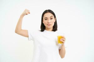 gesund Lebensstil. lächelnd jung asiatisch Frau, zeigt an stark Muskeln, Bizeps auf Hand und Getränke Orange Saft, Weiß Hintergrund foto