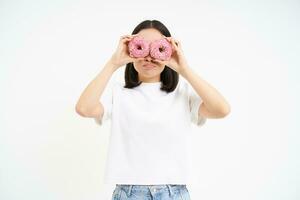 Bild von komisch jung Frau, macht Brille mit zwei Rosa glasiert Donuts, suchen durch Donut Löcher und lächelnd, isoliert auf Weiß Hintergrund foto