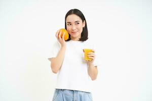 Bild von gesund jung Frau, hält Glas von frisch Saft und Orange Frucht, lächelt nachdenklich, isoliert auf Weiß Hintergrund foto