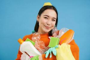 Porträt von begeistert Koreanisch Frau, halten leeren Plastik Flaschen, Sortierung Müll, lächelnd und suchen Glücklich, isoliert auf Blau Hintergrund foto