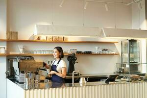 Porträt von Cafe Zähler mit Barista Mädchen Arbeiten mit Kaffee Maschine, Herstellung bestellen zum Klient im Geschäft, tragen Blau Schürze foto