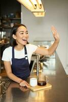 lächelnd asiatisch Mädchen Barista Berufung Klient, geben aus bestellen im Cafe, bereit Stapel brauen Filter Kaffee, tragen Schürze hinter Zähler foto