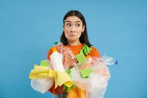 verwirrt asiatisch Frau hält Plastik Abfall, sammelt Müll zu recyceln, lernt Wie zu Sortieren anders Typen von Müll, Konzept von Öko nachhaltig Leben, Blau Hintergrund foto