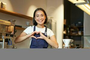 lächelnd asiatisch Mädchen Barista, zeigt an Herz Zeichen, liebt Herstellung Kaffee ein Portion Kunden, Stehen im Uniform hinter Schalter, Arbeit im Cafe foto