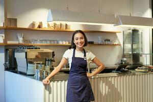 Porträt von süß asiatisch Frau Barista, Cafe Mitarbeiter Stehen in der Nähe von Zähler mit Kaffee Maschine, tragen Schürze, lächelnd beim Kamera foto