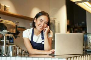 Porträt von lächelnd Koreanisch Frau, Barista im Kaffee Geschäft, Stehen beim Zähler mit Laptop, lächelnd und suchen zuversichtlich, Selbstständiger weiblich Unternehmer im ihr besitzen Kaffee Geschäft foto