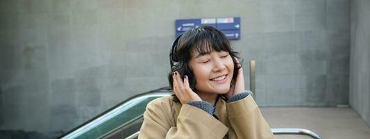 Porträt von glücklich Frau, Koreanisch Mädchen im Kopfhörer, Hören Musik- im Kopfhörer, genießen Klang von Kopfhörer, Lachen und lächelnd foto