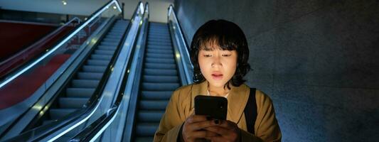 Porträt von asiatisch Mädchen sieht aus schockiert beim Handy, Mobiltelefon Telefon Bildschirm, Stehen auf Rolltreppe, gehen Nieder zu unter Tage oder Metro, pendeln zu Arbeit foto