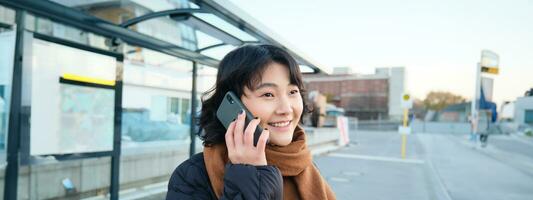 lächeln Koreanisch Mädchen reden auf Handy, Mobiltelefon Telefon, Stehen auf Bus stoppen, mit Smartphone, posieren auf Straße im Winter, eingewickelt im Schal, tragen schwarz Jacke foto