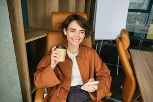 Porträt von weiblich Unternehmer Arbeiten im ihr Büro, halten Smartphone, Trinken Kaffee und lächelnd, genießen ihr Mittagessen brechen foto