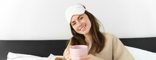 Porträt von schön lächelnd Frau im Schlafen Maske und Pyjama, halten Tasse von Kaffee im Bett, entspannend und suchen glücklich im Bett, aufwachen oben im Morgen foto