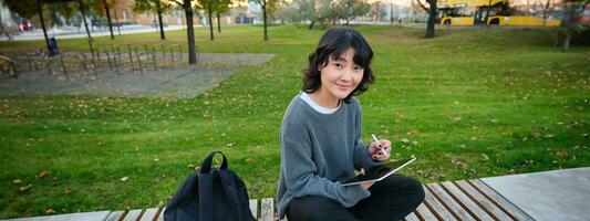 jung asiatisch Mädchen mit Grafik Bleistift und Tablette, sitzt im Park auf Bank, zeichnet Kratzer, tut ihr Hausaufgaben draußen foto