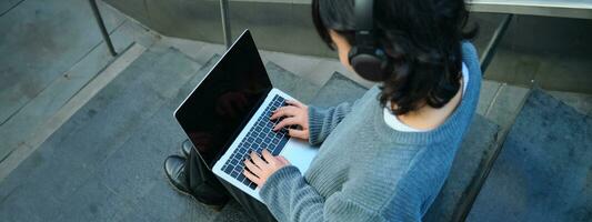 schließen oben Porträt von Mädchen, Schüler funktioniert auf Laptop und hört zu Musik- im Kopfhörer. leer Computer Bildschirm und Hände Tippen auf Tastatur foto