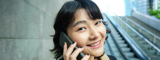 schließen oben Porträt von lächelnd Brünette Mädchen, Koreanisch Frau geht Nieder Rolltreppe, macht Telefon Forderung, Gespräche zu jemand auf Telefon foto