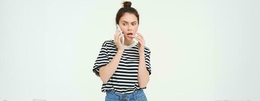 Bild von Frau mit schockiert Gesicht, sieht aus verwirrt während Chats Über das Telefon, hört zu zu Person auf Telefon, Weiß Hintergrund foto