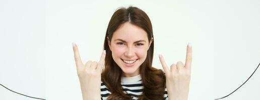 schließen oben Porträt von aufgeregt jung Frau, Felsen n rollen Mädchen, zeigen schwer Metall Horn Finger und lächelnd, genießt Konzert, haben Spaß, Stehen isoliert Über Weiß Hintergrund foto