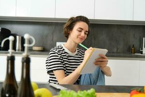 Porträt von schön modern Frau, Mädchen Kochen mit Gemüse, halten Notizbuch, Schreiben Nieder Rezept, Herstellung Anmerkungen, Abendessen Pläne, Erstellen Mahlzeit aufführen zum Wochenende, Sitzung im Küche foto