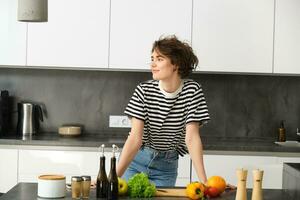 Porträt von jung modern Frau im das Küche, gelehnt auf Zähler mit Hacken Planke, Herstellung ein Salat mit Gemüse und Dressings, Kochen Vegetarier Mahlzeit foto