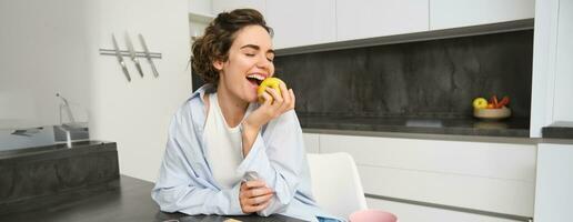 Porträt von glücklich Brünette Frau, beißen ein Apfel, Essen gesund Snack während Sitzung im Küche foto