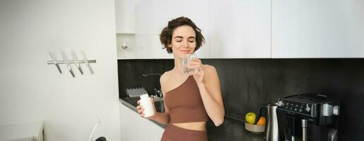 Porträt von Fitness Mädchen, gesund Frau Stehen im Küche mit Pillen, nimmt Vitamine mit Glas von Wasser zum gesund Wohlbefinden. Sportlerin hält Essen Ergänzungen foto