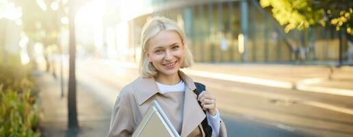 Bild von schön jung lächelnd Frau mit blond Haar, Gehen entlang Straße auf sonnig Tag, tragen ihr Notizbücher und Hochschule Materialien, suchen sorglos foto