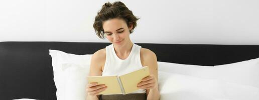 Porträt von Frau lesen ihr Planer und beginnend das Tag, suchen beim Anmerkungen, Sitzung auf Bett im Morgen foto