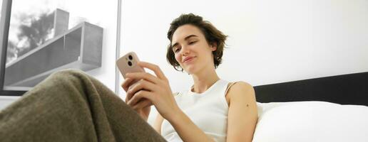 Porträt von jung schön Frau Lügen im Bett, ruhen im Schlafzimmer, Nachrichten, mit Handy, Mobiltelefon Telefon, halten Smartphone und lächelnd, Scrollen Sozial Medien, Ausgaben Komfort Zeit beim Zuhause foto