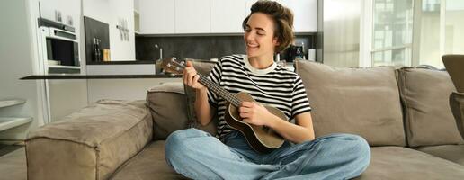 Lebensstil und Hobbys Konzept. jung lächelnd Frau auf Sofa, spielen Ukulele, Singen und Lernen klimpern Muster zum Lieblings Lied foto