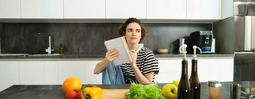 Porträt von jung lächelnd Frau im Küche, halten Notizbuch, Herstellung Anmerkungen zum Rezept, Schreiben Lebensmittelgeschäft Liste, Kochen Salat, Sitzung in der Nähe von Gemüse und Hacken Tafel foto