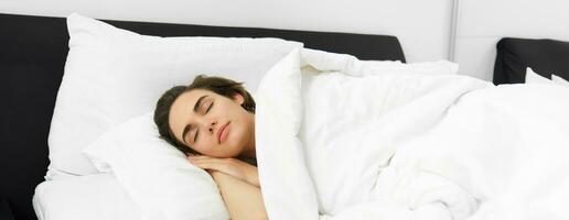 Bild von schön jung Frau Schlafen auf komfortabel Bett, bedeckt mit Sanft Weiß Leinen- Blätter, Augen geschlossen, Gesicht ist Ruhe und ungestört foto