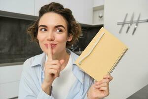 Porträt von süß jung Frau, hält Planer, schreibt ihr Geheimnisse im Tagebuch, zeigt an Stille, shush Geste, hält Finger auf Lippen und sieht aus beiseite, sitzt im Küche foto