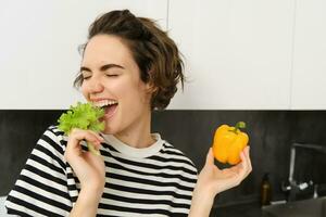 Porträt von sorglos Vegetarier Mädchen, Essen Gemüse, beißen Grüner Salat Blatt mit glücklich lächelnd Gesicht, haben gesund Snack, Likes Gemüse, steht im das Küche foto