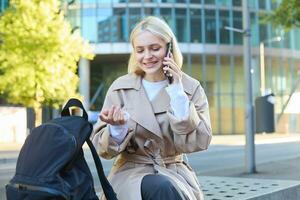 Porträt von jung Frau Sitzung auf Bank, reden auf Handy, Mobiltelefon Telefon, Ausgaben Zeit außen, Antworten ein Telefon Forderung, haben freundlich Konversation auf Handy foto