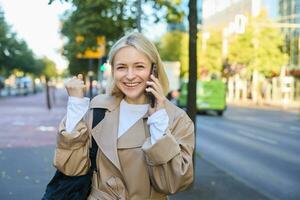 schließen oben Porträt von schön jung Frau, blond Mädchen Gehen auf Straße mit Handy, Mobiltelefon Telefon, chatten mit Freund, hat glücklich Gesicht Ausdruck während reden Über Handy auf ihr Weg foto