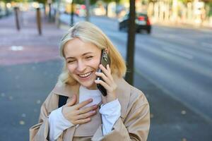 schließen oben Porträt von modern jung Frau, Lachen und lächelnd, Gehen entlang das Straße und Antworten Telefon Forderung, chatten auf Handy, Mobiltelefon Telefon, hat komisch Konversation auf ihr Weg Zuhause foto