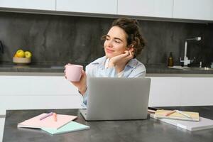 Bild von jung Frau Getränke Tasse von Kaffee und funktioniert von heim. Mädchen Surfen Webseite, Geschäfte online auf Laptop und genießt Tee beim Zuhause foto