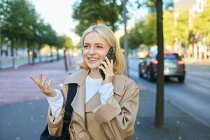 schließen oben Porträt von schön jung Frau, blond Mädchen Gehen auf Straße mit Handy, Mobiltelefon Telefon, chatten mit Freund, hat glücklich Gesicht Ausdruck während reden Über Handy auf ihr Weg foto