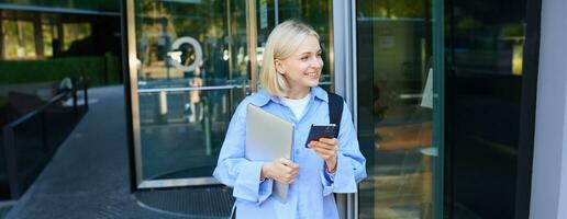 Bild von stilvoll jung Frau, Schüler Gehen aus von das Hochschule Campus, halten Smartphone und Laptop, hat Rucksack auf Schulter, warten zum jemand draußen mit Handy, Mobiltelefon Telefon foto