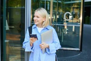 Bild von stilvoll jung Frau, Schüler Gehen aus von das Hochschule Campus, halten Smartphone und Laptop, hat Rucksack auf Schulter, warten zum jemand draußen mit Handy, Mobiltelefon Telefon foto
